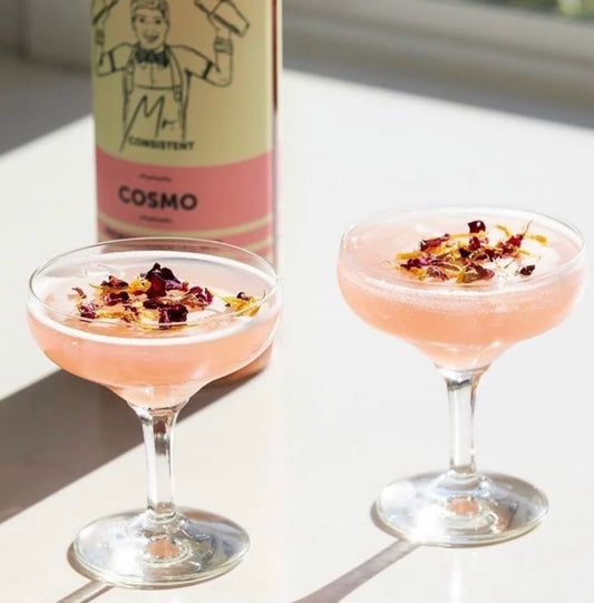 Two pink cocktails in coupe glasses with dried flower petals on top, set on a sunlit white surface, with a bottle labeled COSMO and Concurrent in the background.
