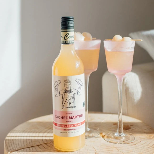 A bottle of Consistent Lychee Martini premium cocktail spirit stands on a wooden table beside two pink cocktails in stemmed glasses, with a softly lit indoor setting in the background.