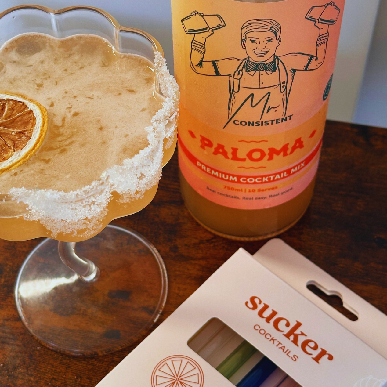 A glass of frothy cocktail with a salted rim and a dried orange slice sits beside a bottle labeled 'Consistent Paloma Premium Cocktail Mix' on a dark wooden table, with a box of 'sucker cocktails' in the foreground.