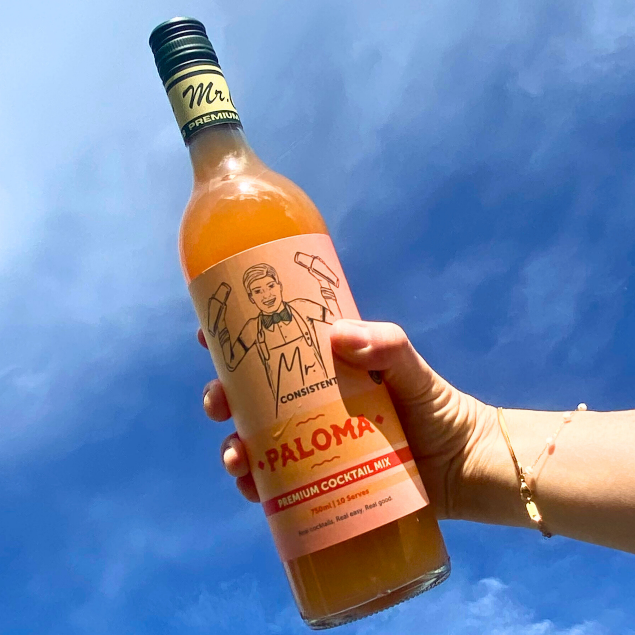 A hand holding a bottle of Mr. Consistant Paloma premium cocktail mix against a bright blue sky background.