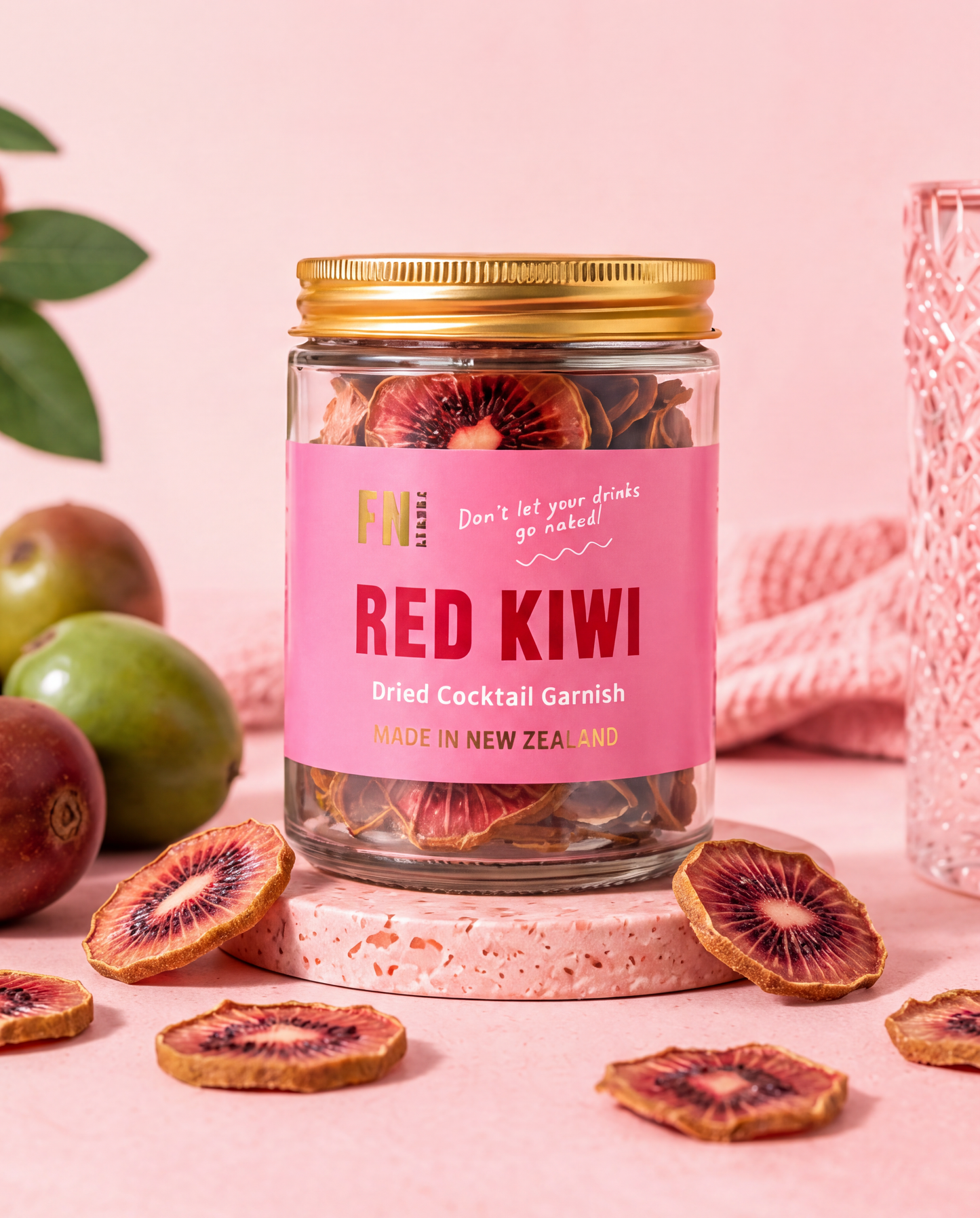 A glass jar of Red Kiwi Dried Cocktail Garnish by F&N Foods, styled on a pink surface with fresh kiwi slices, green plums, and a pink textured glass in the background.