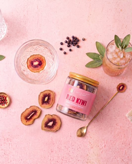 A glass jar labeled 'RED KIWI' with a gold lid and pink label, styled on a pink surface with kiwi slices, a drink in a glass, a spoon, and scattered black seeds.